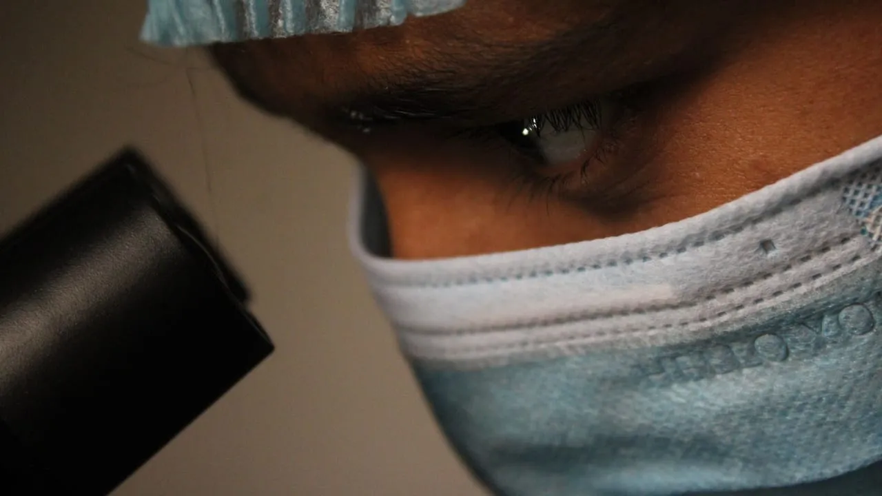 Close-up of a scientist wearing a mask and cap observing through a microscope.