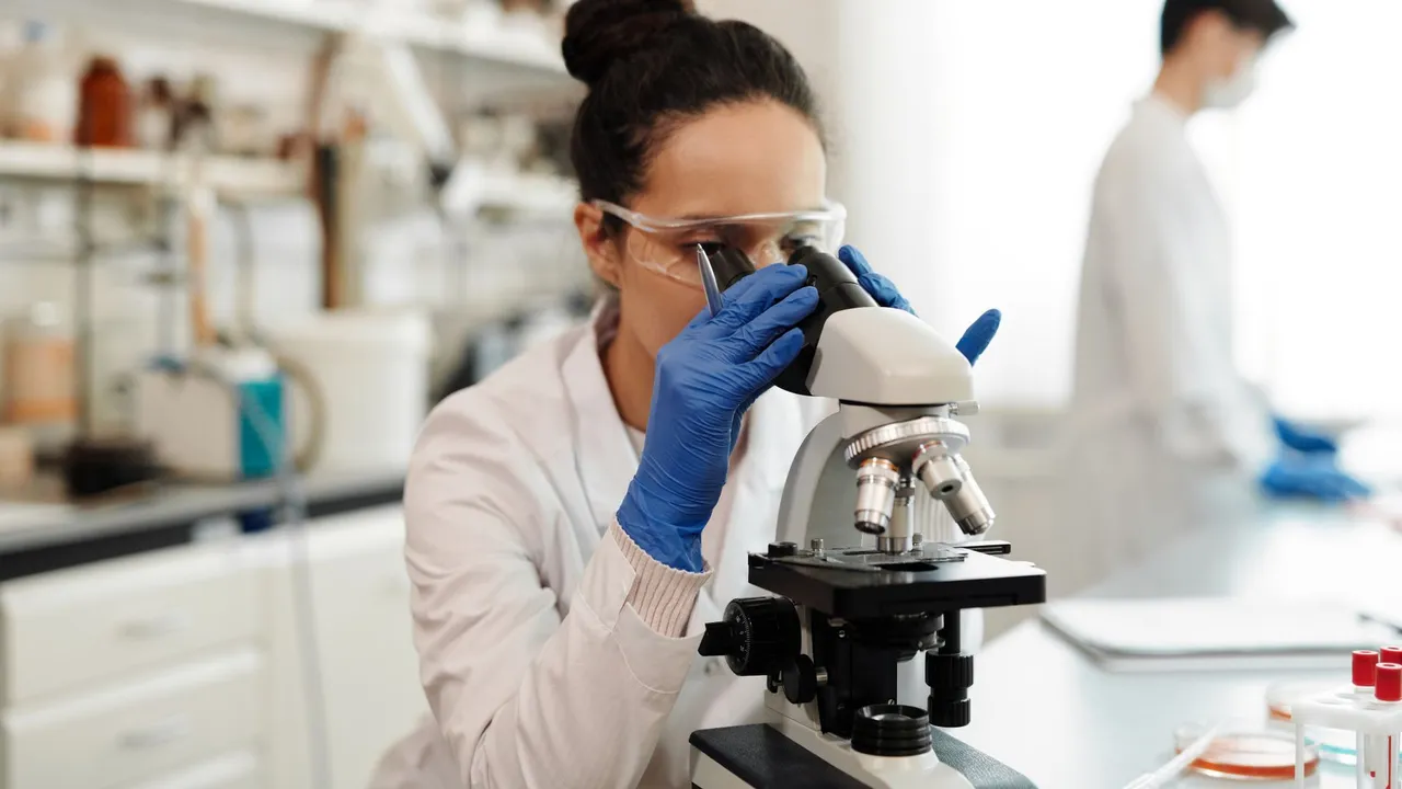A female scientist in protective gear examines samples through a microscope in a laboratory setting.