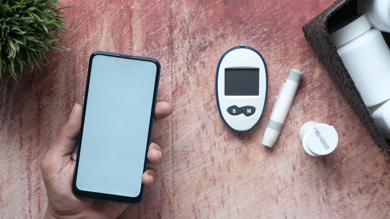 Close-up of a hand holding a smartphone next to a glucometer and medical supplies on a wooden surface.